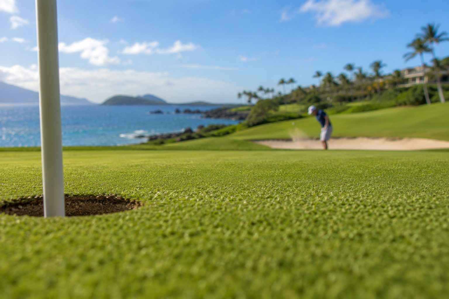 A coastal resort putting green at midday: the flagstick and cup in the foreground, a golfer chipping out of a greenside bunker beyond, with the ocean, a rocky point and palms behind.